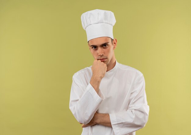 fiducioso giovane maschio cuoco che indossa l'uniforme dello chef mettendo la mano sotto il mento sul muro verde isolato con spazio di copia