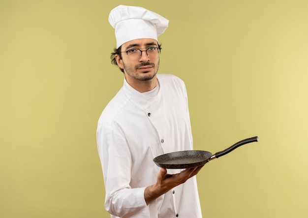 fiducioso giovane maschio cuoco indossando lo chef uniforme e bicchieri tenendo padella su sfondo verde