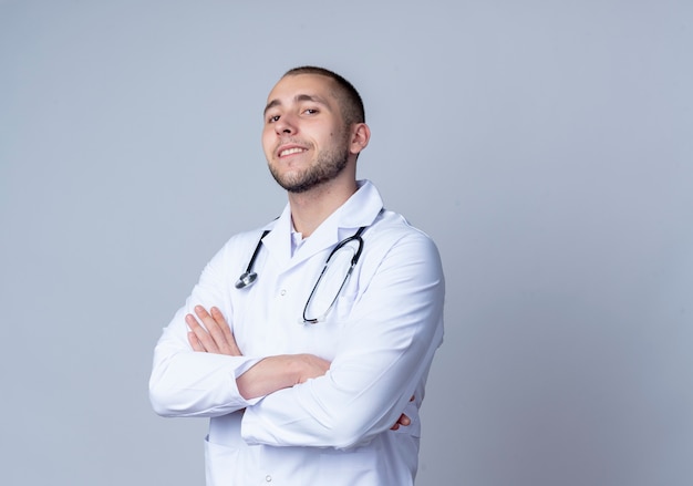 Fiducioso giovane maschio medico indossando abito medico e uno stetoscopio intorno al collo in piedi con la postura chiusa e sorridente isolato su bianco con spazio di copia