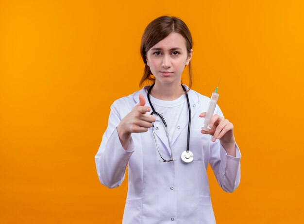 Fiducioso giovane medico femminile che indossa abito medico e stetoscopio tenendo la siringa e ti fa gesto sullo spazio arancione isolato con lo spazio della copia