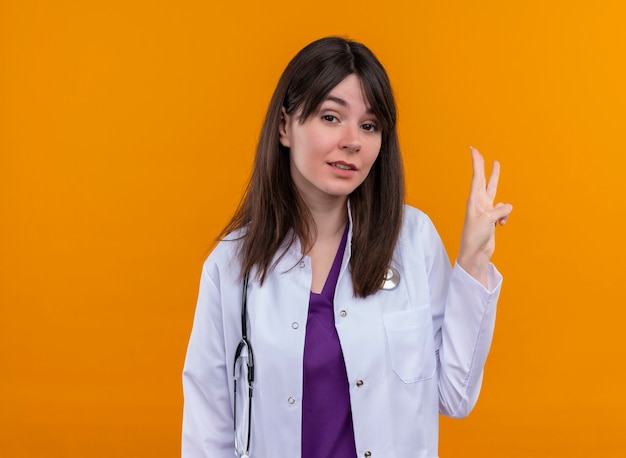 Fiducioso giovane medico femminile in abito medico con lo stetoscopio gesti tre con le dita su sfondo arancione isolato con spazio di copia