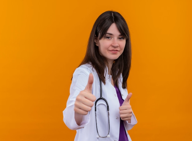 Fiducioso giovane medico femminile in abito medico con lo stetoscopio pollice in alto su sfondo arancione isolato con spazio di copia