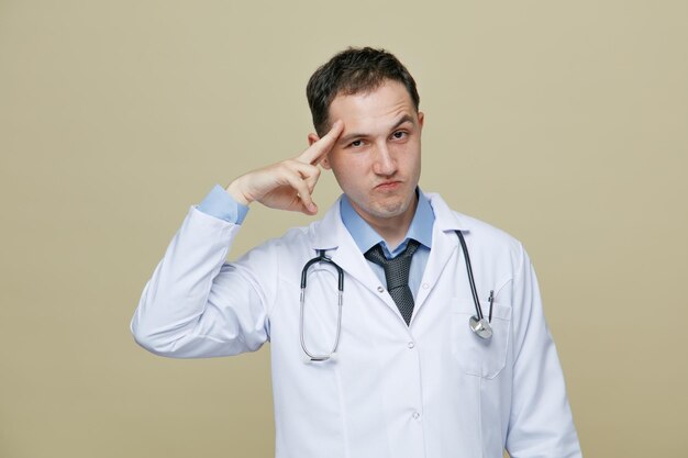 Fiducioso giovane medico maschio che indossa accappatoio medico e stetoscopio intorno al collo guardando la fotocamera facendo gesto di saluto isolato su sfondo verde oliva