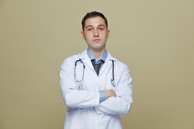 fiducioso giovane medico maschio che indossa un abito medico e uno stetoscopio intorno al collo guardando la fotocamera tenendo le braccia incrociate isolate su sfondo verde oliva