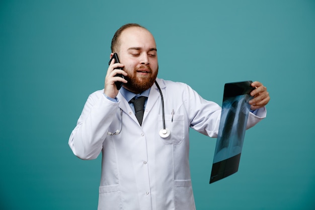 Fiducioso giovane medico maschio che indossa un camice medico e uno stetoscopio intorno al collo che tiene e guarda i raggi x mentre parla al telefono isolato su sfondo blu