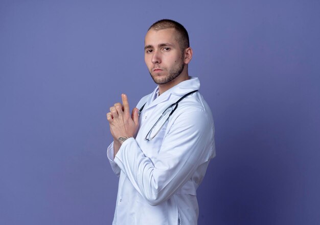 Fiducioso giovane medico maschio che indossa veste medica e stetoscopio intorno al collo in piedi in vista di profilo facendo gesto di pistola con la mano e guardando davanti isolato sul muro viola