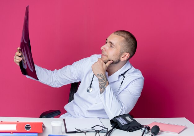 Fiducioso giovane medico maschio che indossa veste medica e stetoscopio seduto alla scrivania con strumenti di lavoro che tengono e guardando i raggi x girato con la mano sul mento isolato sul rosa