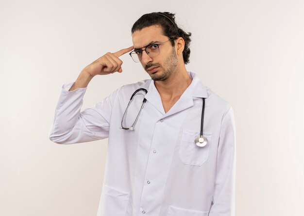 Fiducioso giovane medico maschio con occhiali ottici che indossa una veste bianca con lo stetoscopio che mette il dito sulla fronte