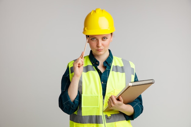 Fiducioso giovane operaio edile femminile che indossa il casco di sicurezza e il blocchetto per appunti della tenuta della maglia di sicurezza che fa pensare il gesto con la matita