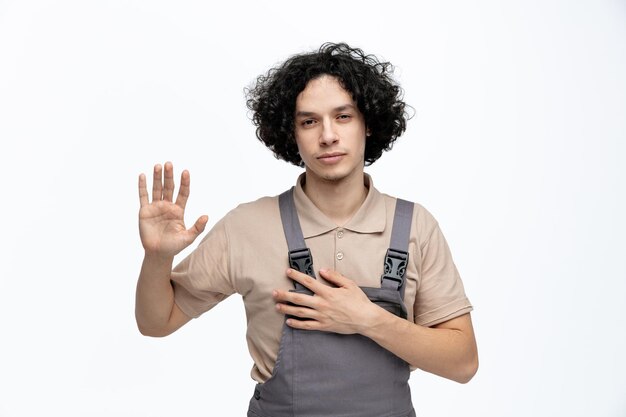 Fiducioso giovane operaio edile maschio che indossa l'uniforme guardando la fotocamera tenendo la mano sul petto che mostra la mano vuota isolata su sfondo bianco