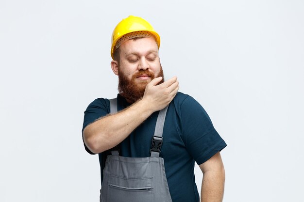 Fiducioso giovane operaio edile maschio che indossa un casco di sicurezza e uniforme spolverare la spalla guardando la sua spalla isolata su sfondo bianco