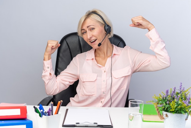 Fiducioso giovane operatore di call center femminile che indossa l'auricolare seduto al tavolo con strumenti da ufficio facendo un gesto forte isolato sul muro bianco