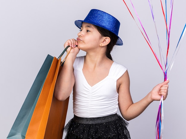 Fiducioso giovane ragazza caucasica con cappello da festa blu che tiene palloncini di elio e borse della spesa guardando il lato isolato sul muro bianco con spazio di copia