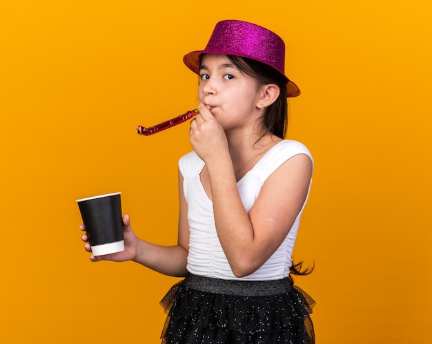 Fiducioso giovane ragazza caucasica con viola party hat tenendo il bicchiere di carta e soffiando il fischio del partito isolato sulla parete arancione con spazio di copia