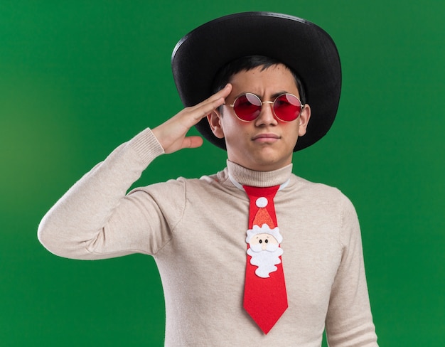 Fiducioso giovane ragazzo che indossa il cappello con cravatta natalizia e occhiali che mostrano gesto di saluto isolato sulla parete verde