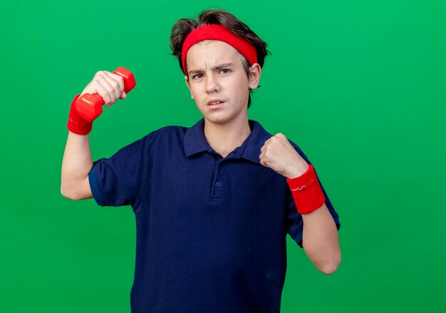 Fiducioso giovane ragazzo sportivo bello che indossa fascia e braccialetti con bretelle dentali alzando il pugno di serraggio con manubri guardando davanti isolato sulla parete verde con lo spazio della copia