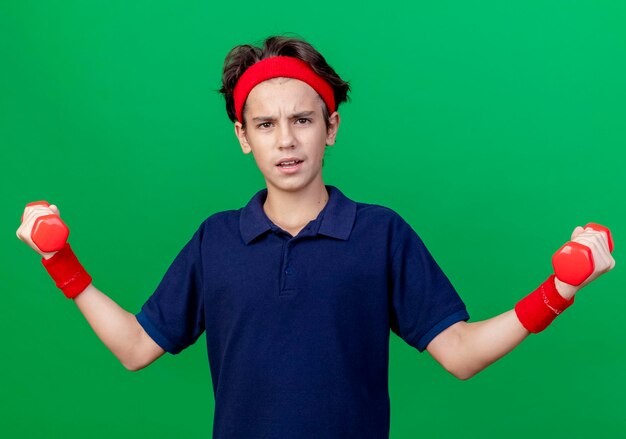 Fiducioso giovane ragazzo sportivo bello che indossa la fascia e braccialetti con bretelle dentali che tengono i manubri guardando la parte anteriore isolata sulla parete verde