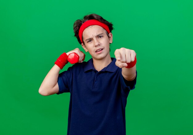 Fiducioso giovane ragazzo sportivo bello che indossa la fascia e braccialetti con bretelle dentali che tengono il manubrio guardando e indicando davanti isolato sulla parete verde con lo spazio della copia