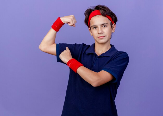 Fiducioso giovane ragazzo sportivo bello che indossa la fascia e braccialetti con le parentesi graffe dentali facendo un forte gesto che indica i suoi muscoli isolati sulla parete viola con lo spazio della copia