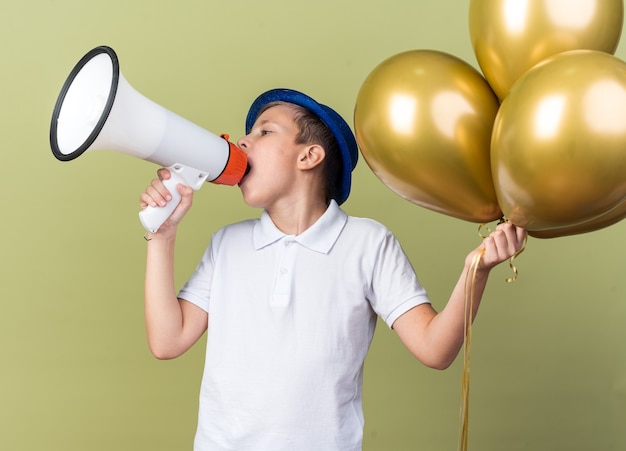 fiducioso, giovane, slavo, ragazzo, con, blu, festa, cappello, presa a terra, elio, palloni, e, parlare, in, altoparlante, guardando, lato, isolato, su, verde oliva, parete, con, spazio copia