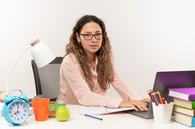 Fiducioso giovane studentessa graziosa con gli occhiali seduto alla scrivania con strumenti di scuola facendo i compiti utilizzando laptop isolato su bianco