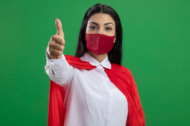 Fiducioso giovane superdonna che indossa la maschera guardando la parte anteriore che mostra pollice in alto isolato sulla parete verde