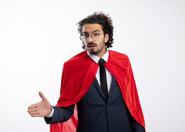 Fiducioso giovane supereroe caucasico uomo in occhiali ottici che indossa un abito con mantello rosso tiene la mano guardando la telecamera