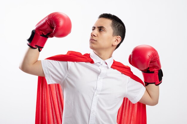 Fiducioso giovane supereroe ragazzo in mantello rosso indossando guanti boxe facendo un forte gesto guardando la sua mano isolata su sfondo bianco