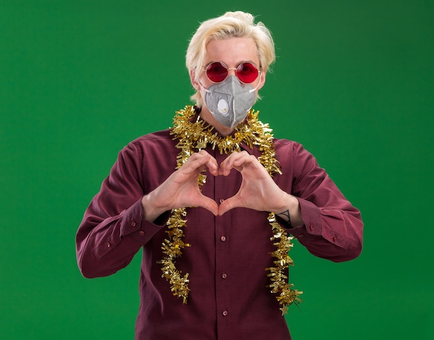 Fiducioso giovane uomo biondo con gli occhiali e maschera protettiva con tinsel ghirlanda intorno al collo guardando la telecamera facendo segno di cuore isolato su sfondo verde