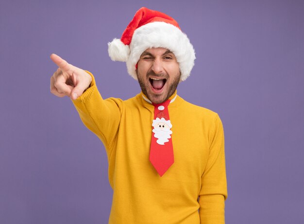 Fiducioso giovane uomo caucasico che indossa cappello di Natale e cravatta guardando la telecamera sbattere le palpebre rivolto a lato con la bocca aperta isolato su sfondo viola con spazio di copia