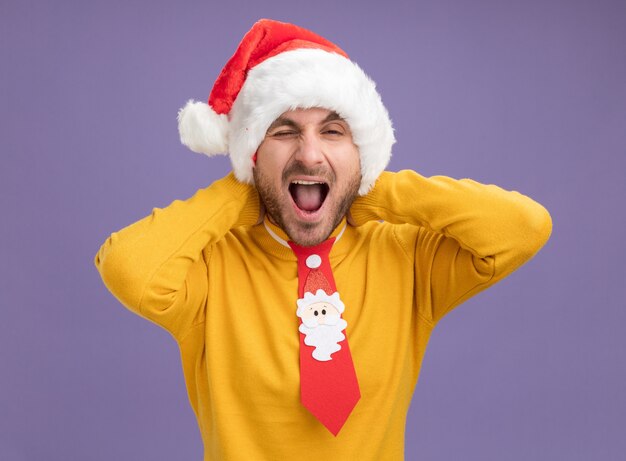 Fiducioso giovane uomo caucasico che indossa il cappello di Natale e cravatta tenendo le mani dietro il collo guardando la telecamera sbattere le palpebre con la bocca aperta isolata su sfondo viola