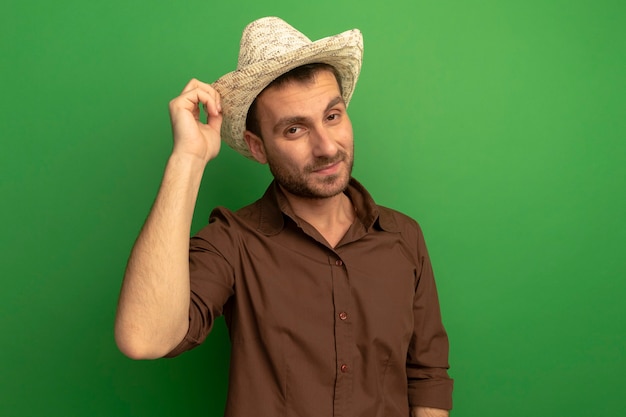 Fiducioso giovane uomo che indossa cappello da spiaggia afferrandolo guardando davanti isolato sul muro verde