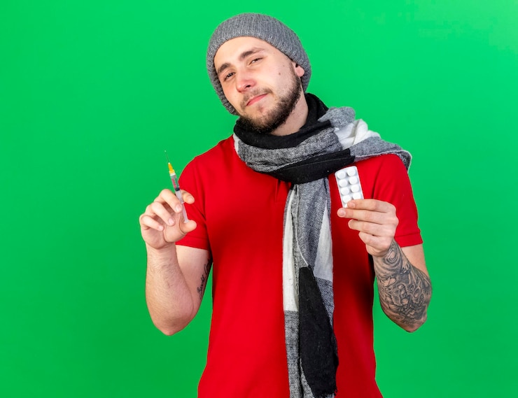 Foto gratuito fiducioso giovane uomo malato caucasico che indossa sciarpa e cappello invernale tiene la siringa e il pacchetto di pillole mediche sul verde