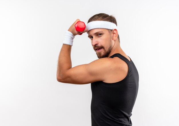 Fiducioso giovane uomo sportivo bello che indossa fascia e braccialetti che alzano il manubrio isolato sul muro bianco con copia spazio