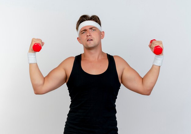 Fiducioso giovane uomo sportivo bello indossando la fascia e braccialetti alzando manubri isolati su bianco