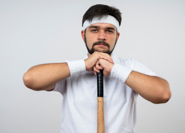 Fiducioso giovane uomo sportivo che indossa la fascia e il braccialetto che mette il mento sulla mazza da baseball isolato sul muro bianco