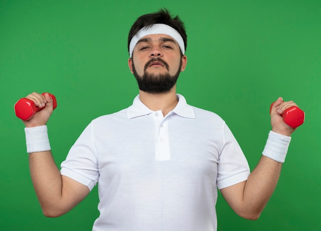 Fiducioso giovane uomo sportivo indossa la fascia e il braccialetto che si esercita con i dumbbells isolati sul verde