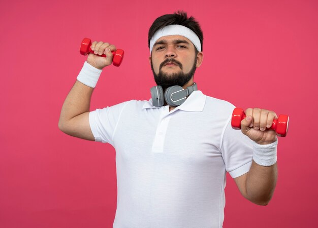 Fiducioso giovane uomo sportivo indossa la fascia e il braccialetto con le cuffie che si esercitano con i manubri