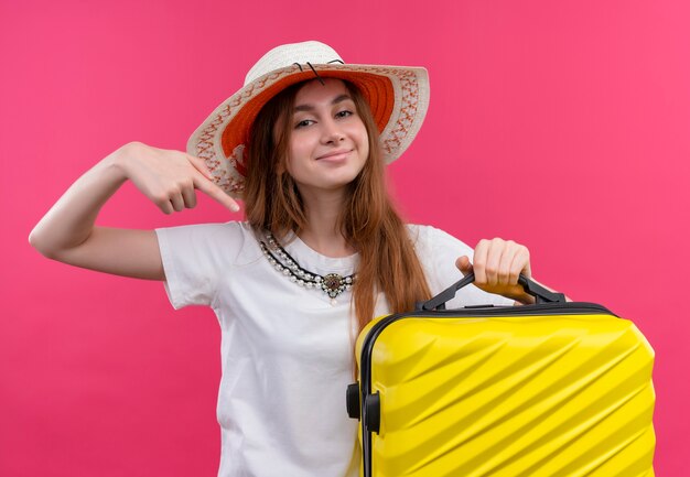 Fiducioso giovane viaggiatore ragazza che indossa il cappello tenendo la valigia e indicandolo su uno spazio rosa isolato