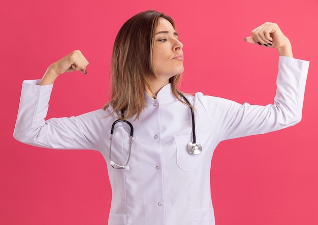 Fiducioso guardando il lato giovane dottoressa che indossa un abito medico con uno stetoscopio che mostra un gesto forte isolato sulla parete rosa pink