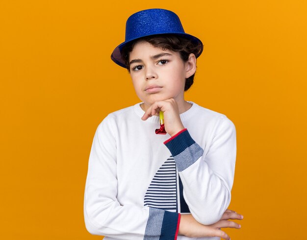 Fiducioso ragazzino che indossa il cappello da festa blu che tiene il fischio della festa mettendo la mano sotto il mento isolato sulla parete arancione