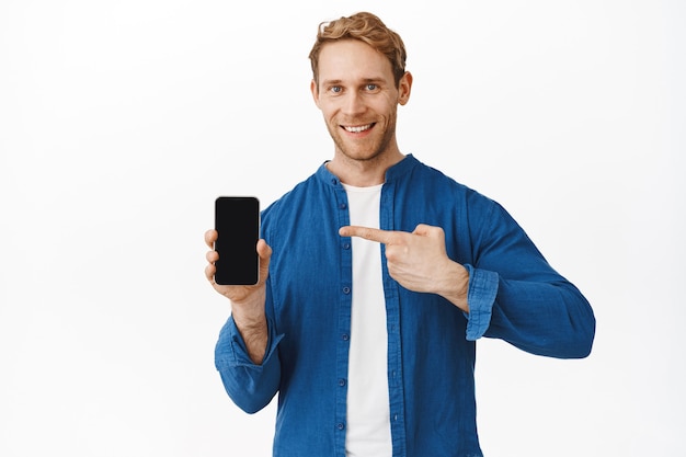 Fiducioso sorridente uomo rosso che punta allo schermo del telefono e guarda davanti, consiglia una buona applicazione su smartphone, mostra affare, muro bianco