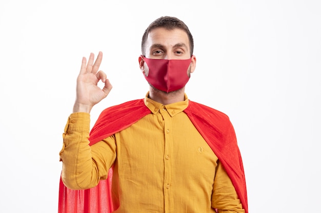 Fiducioso supereroe uomo con mantello rosso che indossa la maschera rossa gesti segno ok con la mano guardando la parte anteriore isolata sul muro bianco