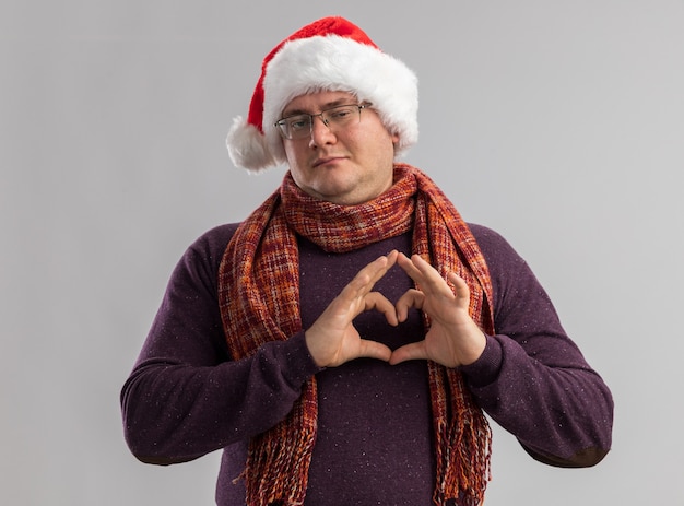 Fiducioso uomo adulto con gli occhiali e cappello da Babbo Natale con sciarpa intorno al collo facendo segno di cuore isolato su muro bianco