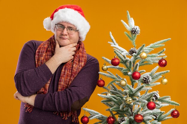 Fiducioso uomo adulto con gli occhiali e cappello da Babbo Natale con sciarpa intorno al collo in piedi vicino all'albero di Natale decorato tenendo la mano sul mento isolato sul muro arancione