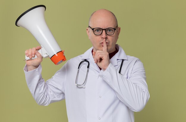 Fiducioso uomo adulto con gli occhiali in uniforme da medico con stetoscopio che tiene in mano un altoparlante e fa il gesto del silenzio