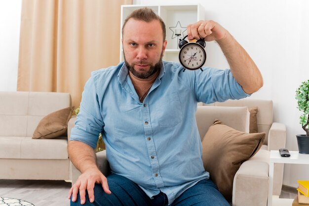 Fiducioso uomo adulto slavo si siede sulla poltrona tenendo sveglia all'interno del soggiorno