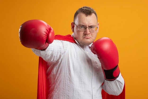 Fiducioso uomo adulto supereroe in mantello rosso con gli occhiali e guanti di scatola guardando davanti facendo gesto di boxe isolato sulla parete arancione