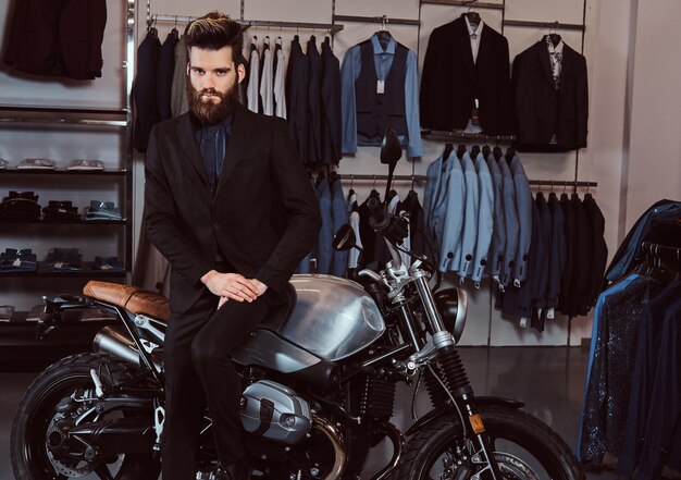 Fiducioso uomo alla moda con la barba vestito in abito nero appoggiato su una moto sportiva retrò nel negozio di abbigliamento maschile.