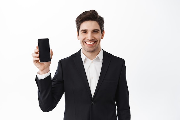 Fiducioso uomo d'affari in abito nero che mostra lo schermo dello smartphone vuoto, sorridente, presenta un'applicazione, un negozio online o un promo, sfondo bianco